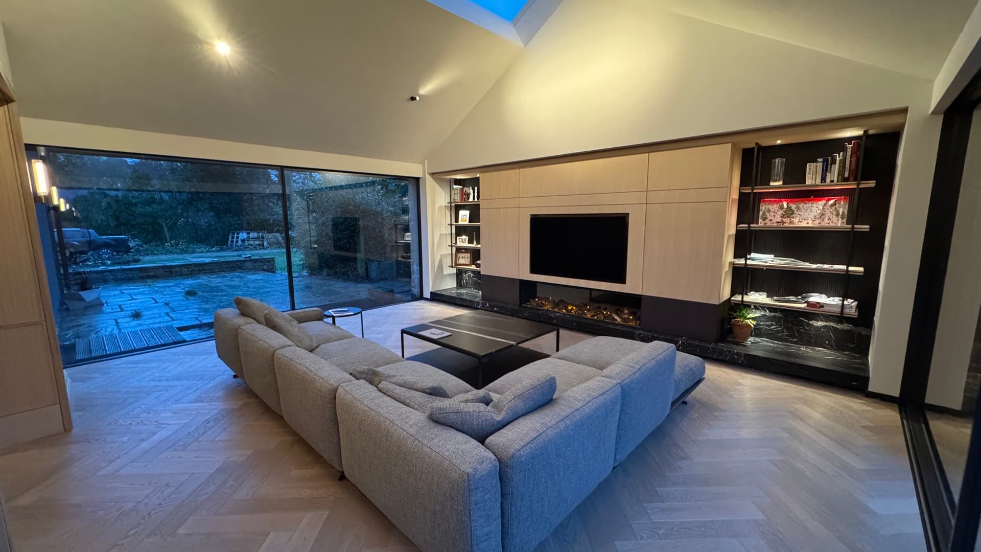 An open-plan living room with a media wall, concealed lighting and a linear fire, opening onto a garden terrace