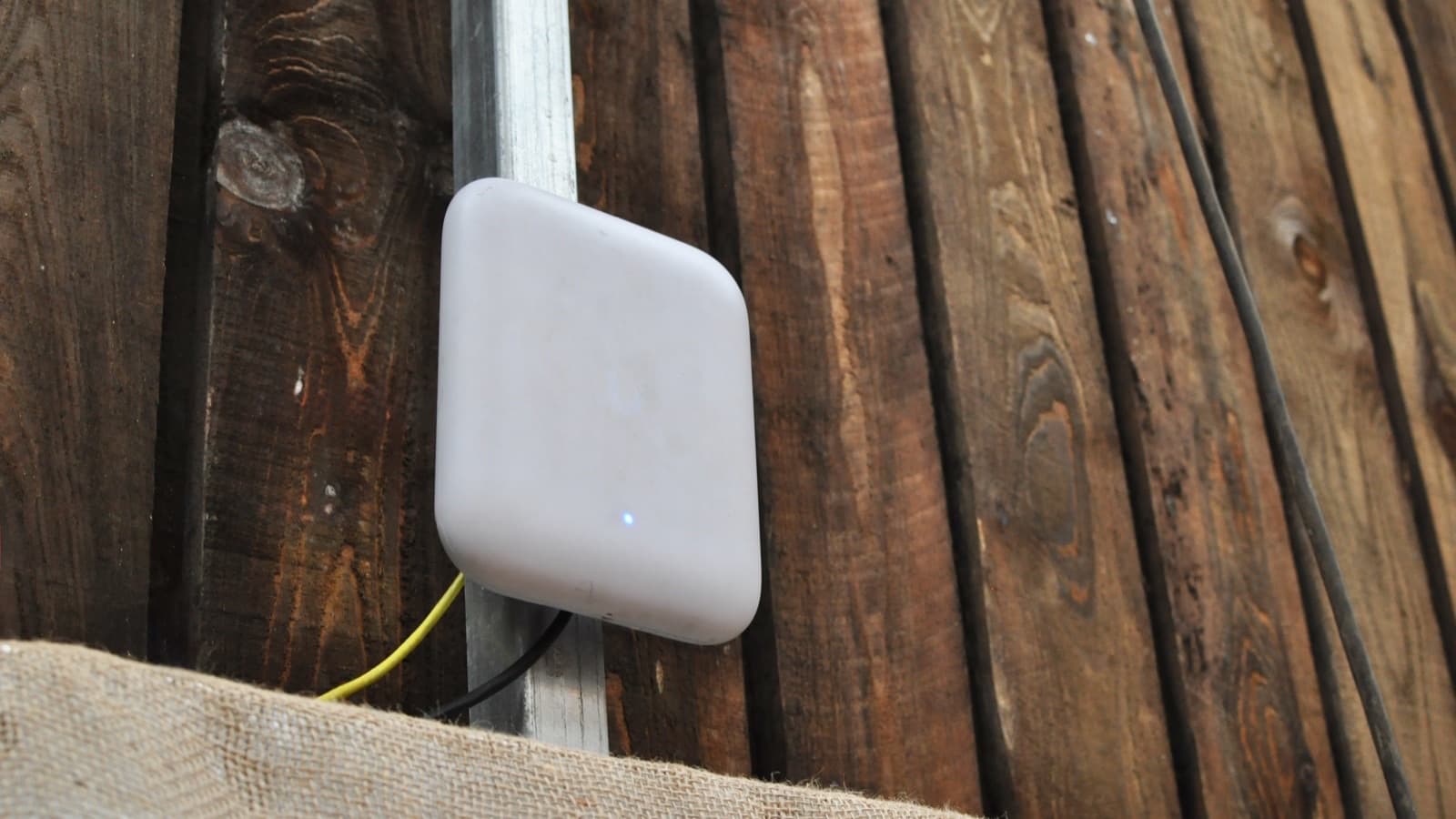 An outdoor wireless access point mounted to a steel rail against timber boarding, its status light lit